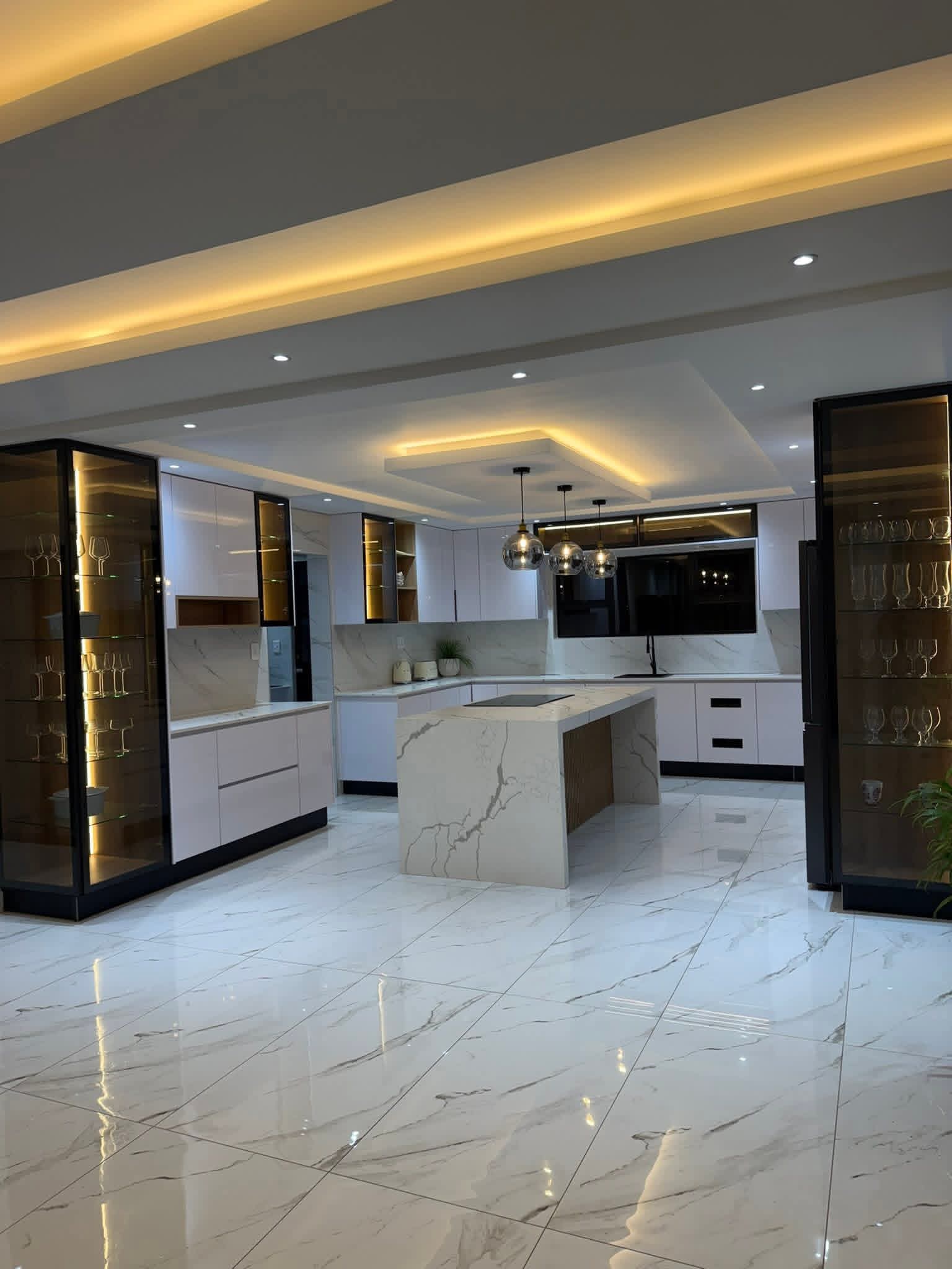 Wide kitchen with layered tray ceilings, cove lighting, marble floors and twin glass display towers